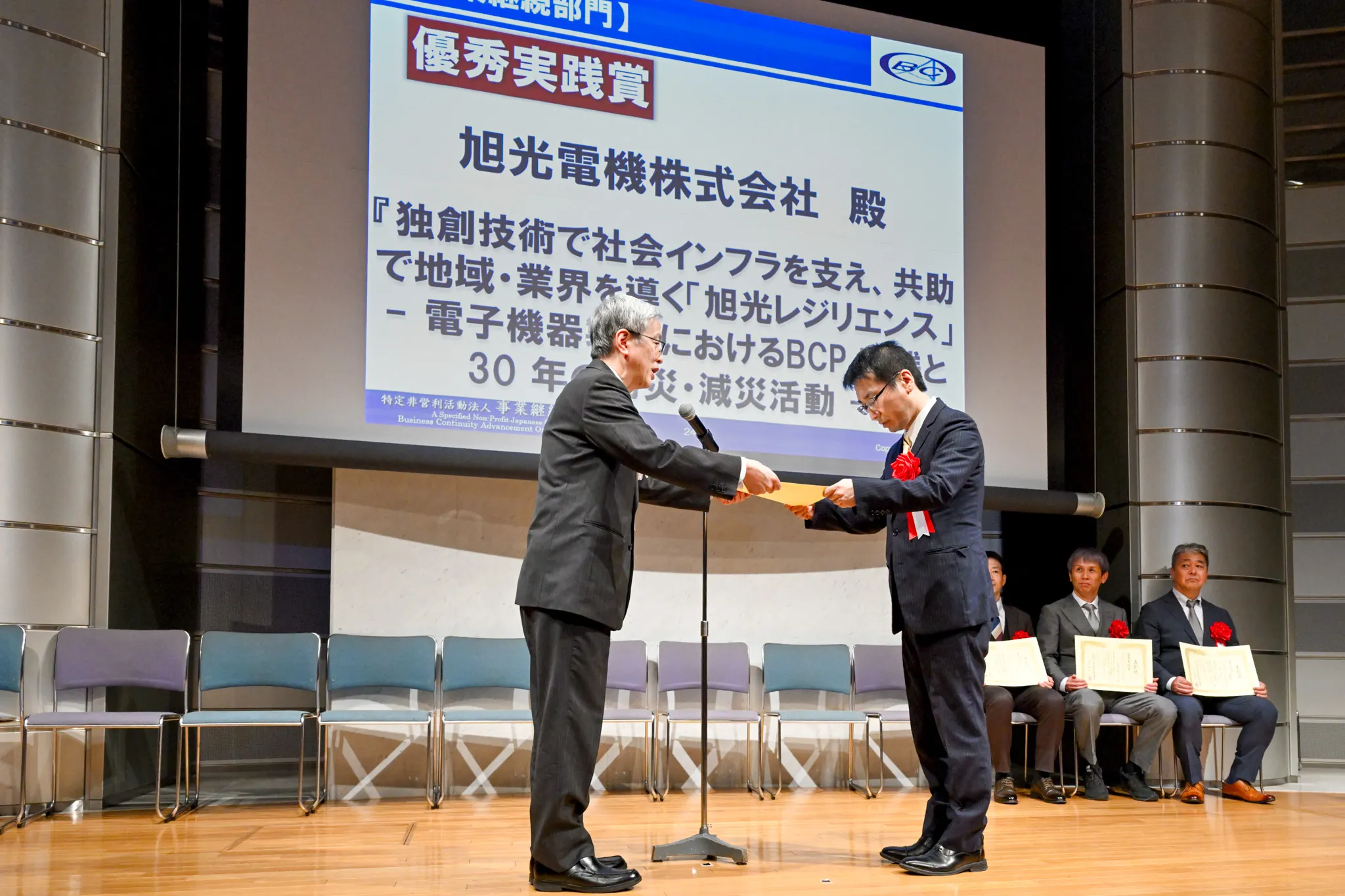 BCAOアワード2025表彰式で、旭光電機株式会社が『優秀実践賞』を受賞し、壇上で表彰状を授与されている様子