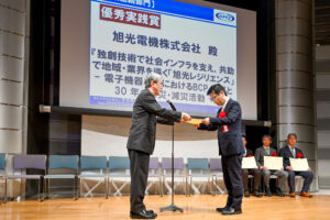BCAOアワード2025表彰式で、旭光電機株式会社が『優秀実践賞』を受賞し、壇上で表彰状を授与されている様子