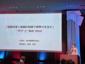 DXセレクション2026の最終選考の場で、スクリーンに資料を投影しながら旭光電機のDX推進の取り組みを発表する社長