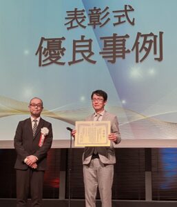 DXセレクション2026の表彰式で『優良事例』として表彰され、賞状を受け取り掲げる社長の様子